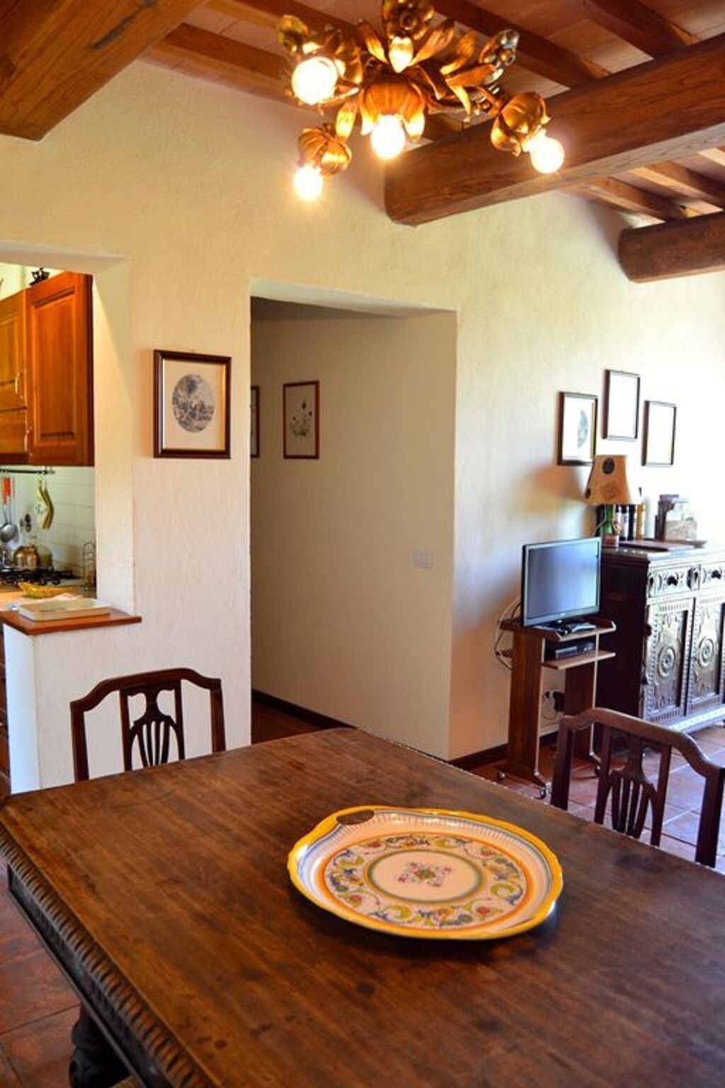 A dining area with a wooden table, chairs, and a decorative plate. There is a television on a stand and a cabinet with a lamp.