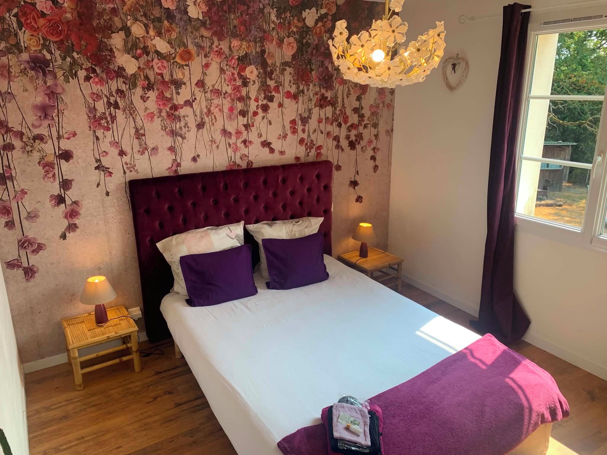 A bedroom with a large bed, two bedside lamps, a hanging floral arrangement, and a window with curtains.