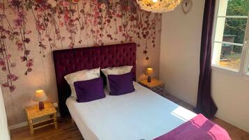 A bedroom with a large bed, two bedside lamps, a hanging floral arrangement, and a window with curtains.