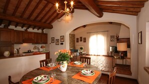 A dining area with a round table set for four, surrounded by wooden chairs. A chandelier hangs from the ceiling, and a vase with flowers is placed on the table.