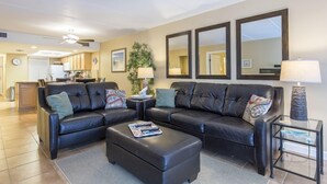 A living room with a black leather sofa, a matching ottoman, and a glass coffee table. There are three large mirrors on the wall, a potted plant, and a lamp on a side table.