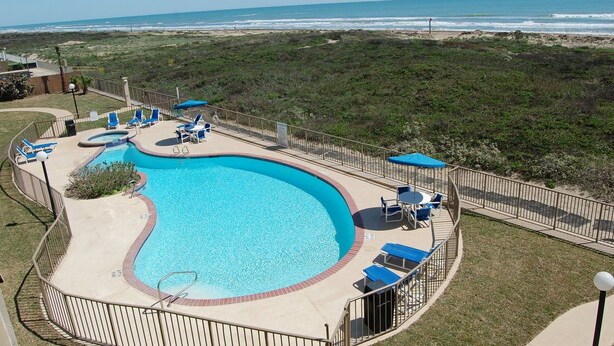 A pool area with a curved design, surrounded by a fence, and overlooking a beach.
