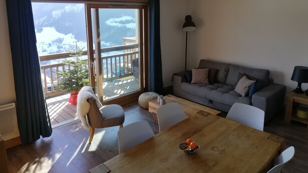 Uma sala de estar moderna com uma mesa de jantar de madeira, sofá cinza e vista para montanhas nevadas através de uma grande porta de vidro deslizante.
