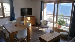 Uma sala de estar moderna com uma mesa de jantar de madeira, cadeiras brancas, um sofá, uma TV e uma varanda com vista para montanhas.