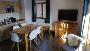 Cozinha moderna com micro-ondas, torradeira e pia. Há uma mesa de jantar com cadeiras e uma televisão sobre um armário de madeira.