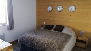 Quarto com cabeceira de madeira, três relógios de parede, cama com manta fofa, abajures e janela com cortinas.