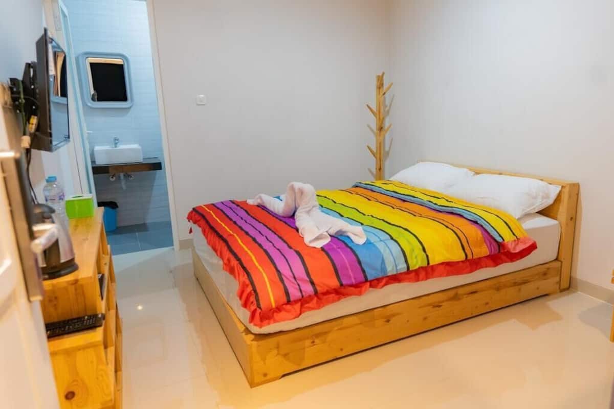 A single bed with a colorful striped blanket, a wooden headboard, and a white pillow. A wooden nightstand with a lamp and a bottle of water is to the left. A small window with a view of a bathroom is visible in the background.