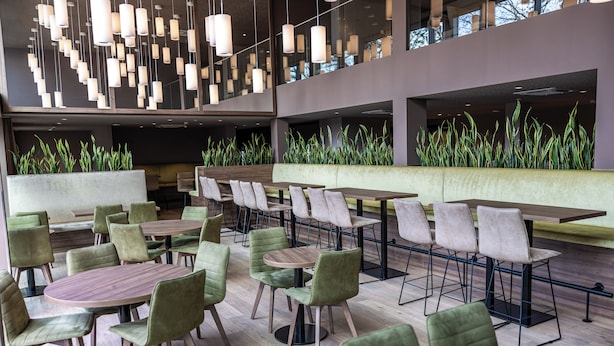 Un restaurante moderno con mesas redondas y sillas verdes, cuenta con un techo alto con luces colgantes y vistas de la ciudad a través de amplias ventanas.