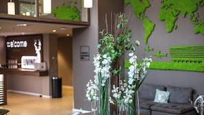 Un área de recepción con un cartel que dice 'Welcome', otro cartel que dice 'Living' y un mapa del mundo en la pared. Hay jarrones con flores blancas sobre una mesa.