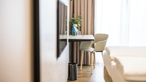 Una habitación de hotel con cama, escritorio, silla, un cesto de basura pequeño y ventana con cortinas.