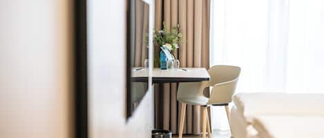 A hotel room with a bed, a desk with a chair, a small trash bin, and a window with curtains.