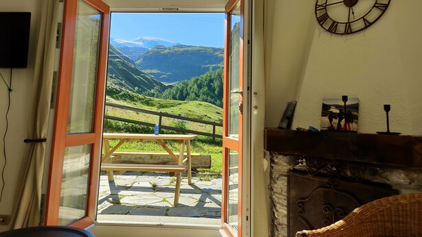 Une porte ouverte donne sur un patio équipé d’une table et de bancs en bois, ainsi que d’un sol en pierre. On y aperçoit une horloge au mur et une vue sur un paysage montagneux.