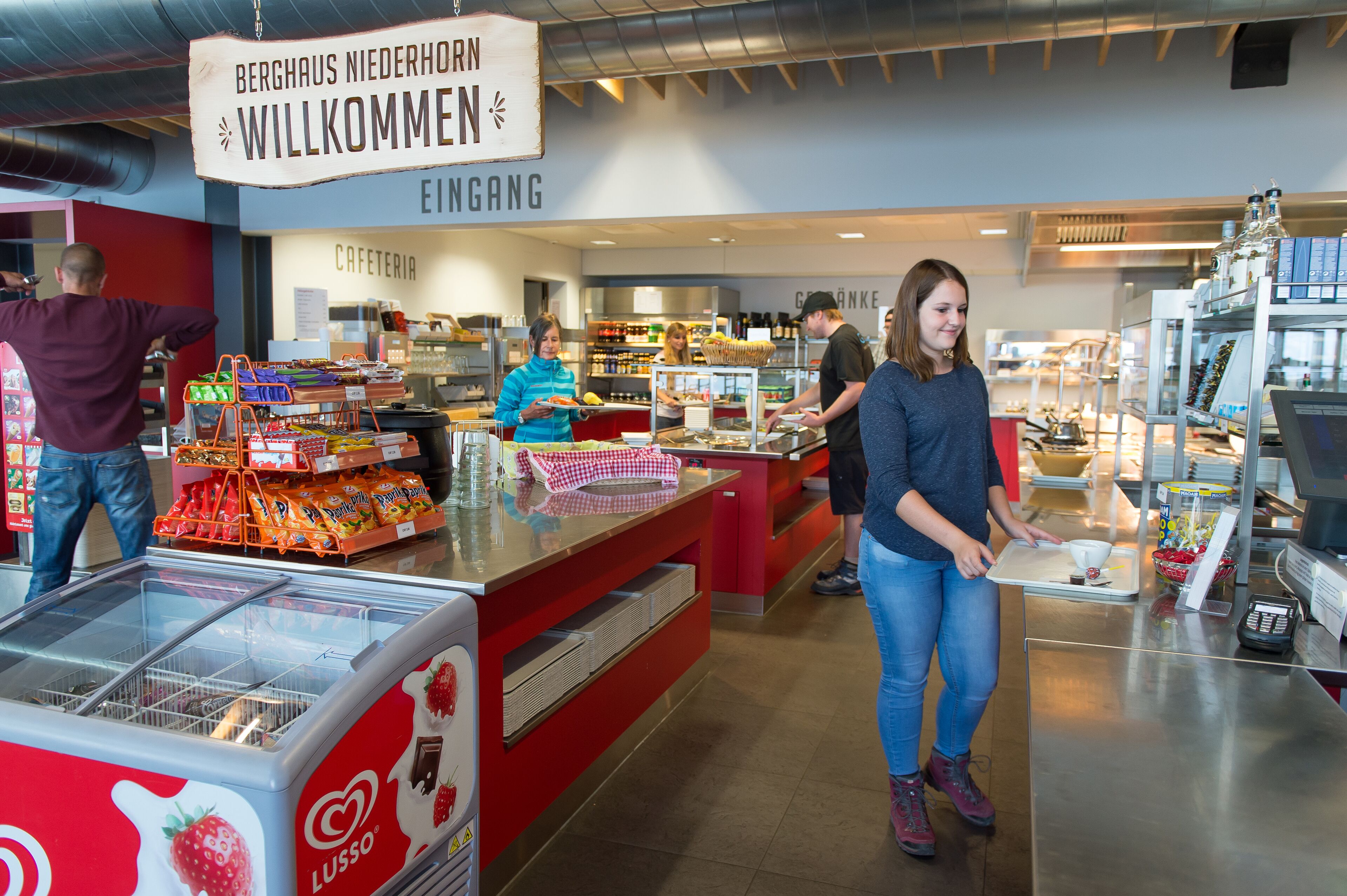 A cafeteria area with a sign reading "Berghaus Niederhorn Willkommen" and a counter with food items.
