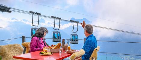 A dining table with food and a beverage, a cable car in the background, and a mountainous landscape.
