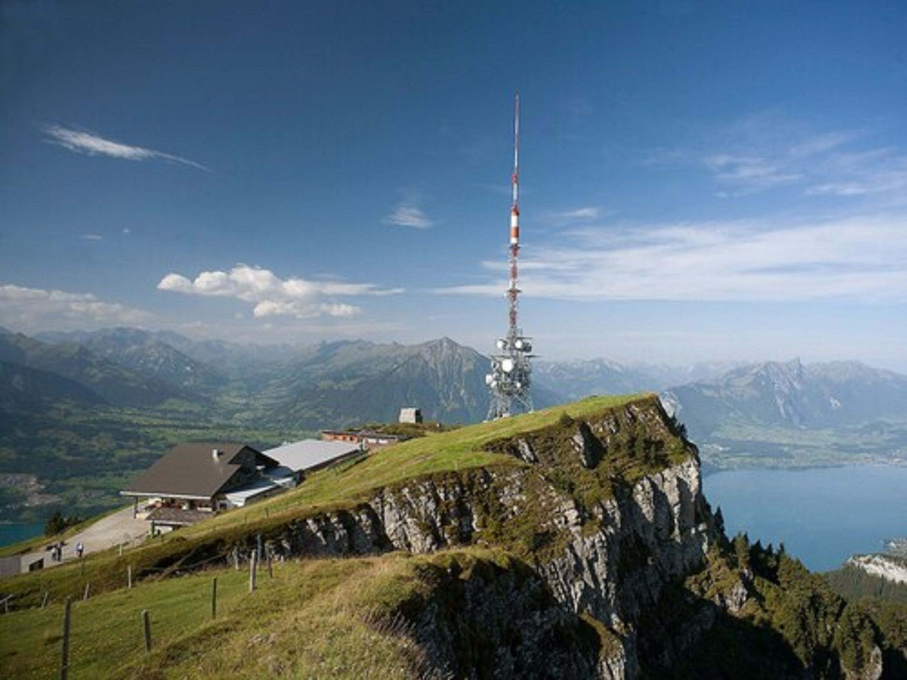 A mountain lodge with a prominent tower, surrounded by lush greenery and a lake.