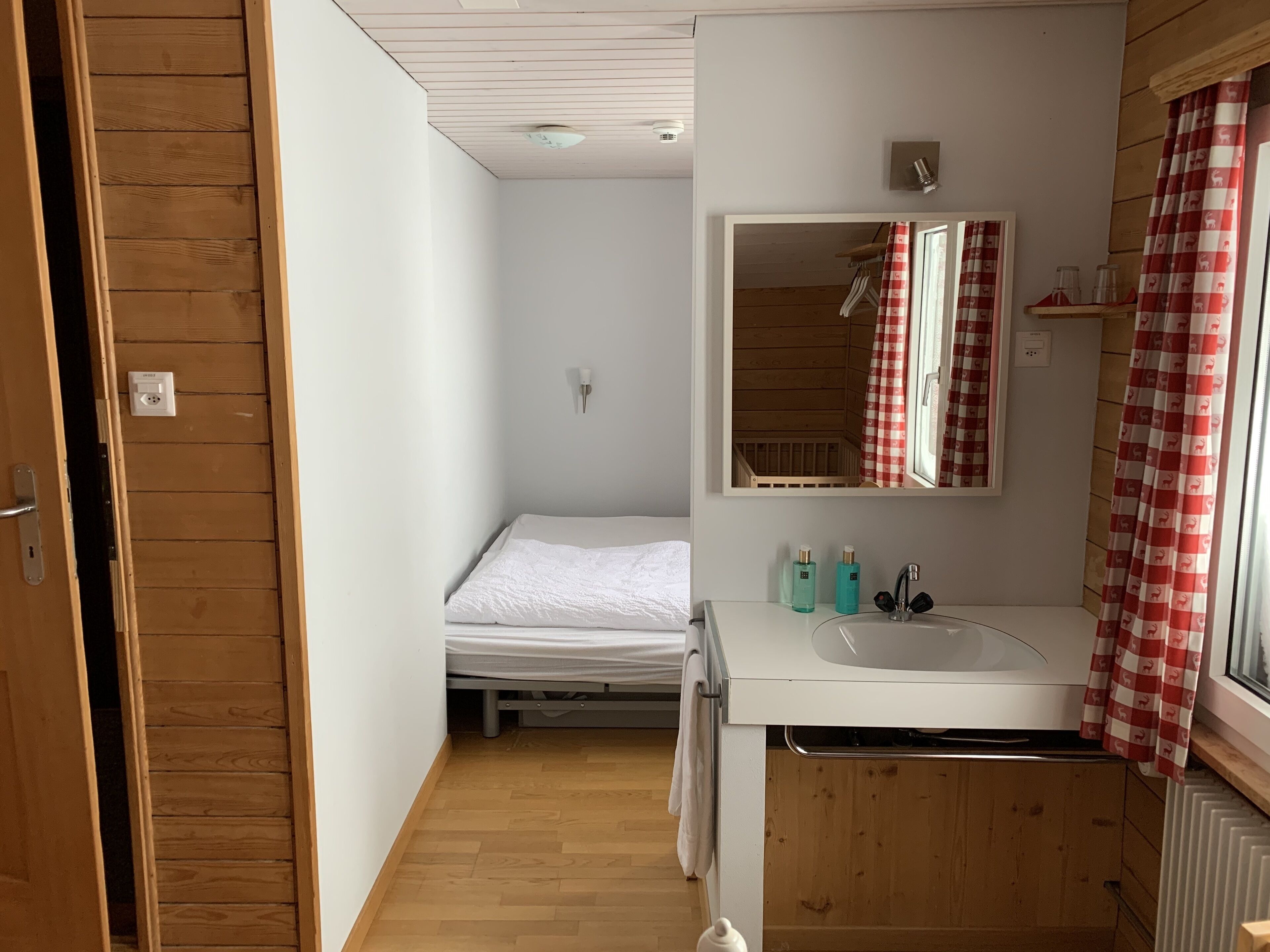 A compact bedroom with a single bed, a small bathroom with a sink and mirror, and wooden walls.