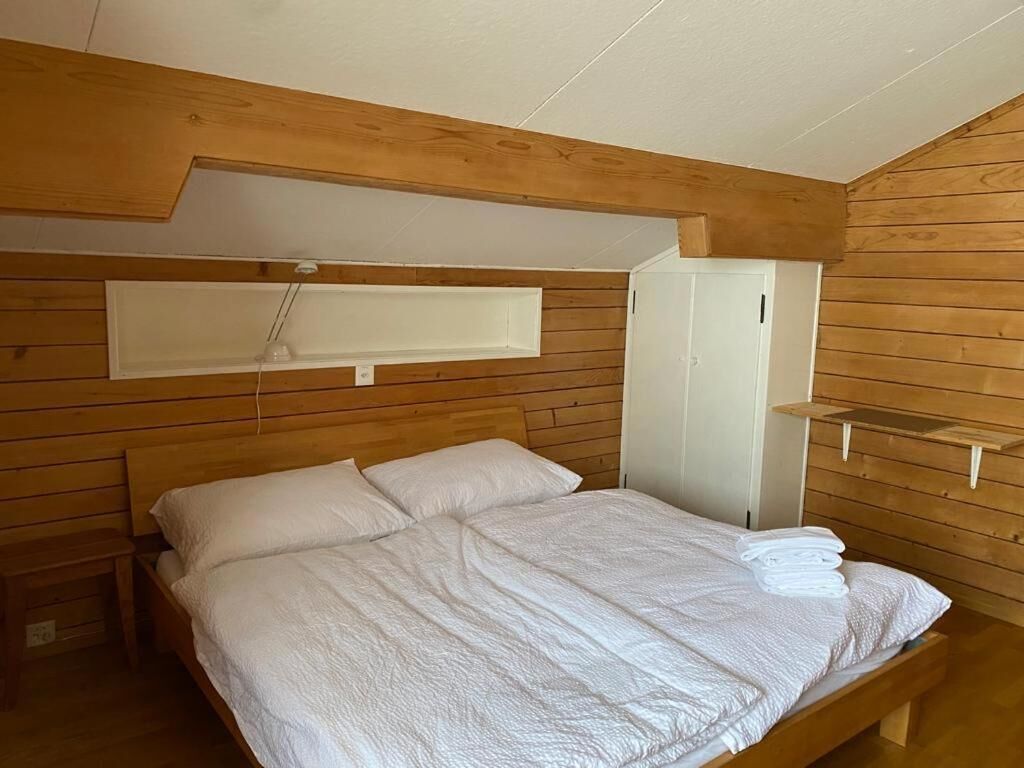 A bedroom with a wooden ceiling, a bed with white bedding, a white wardrobe, and a small shelf.