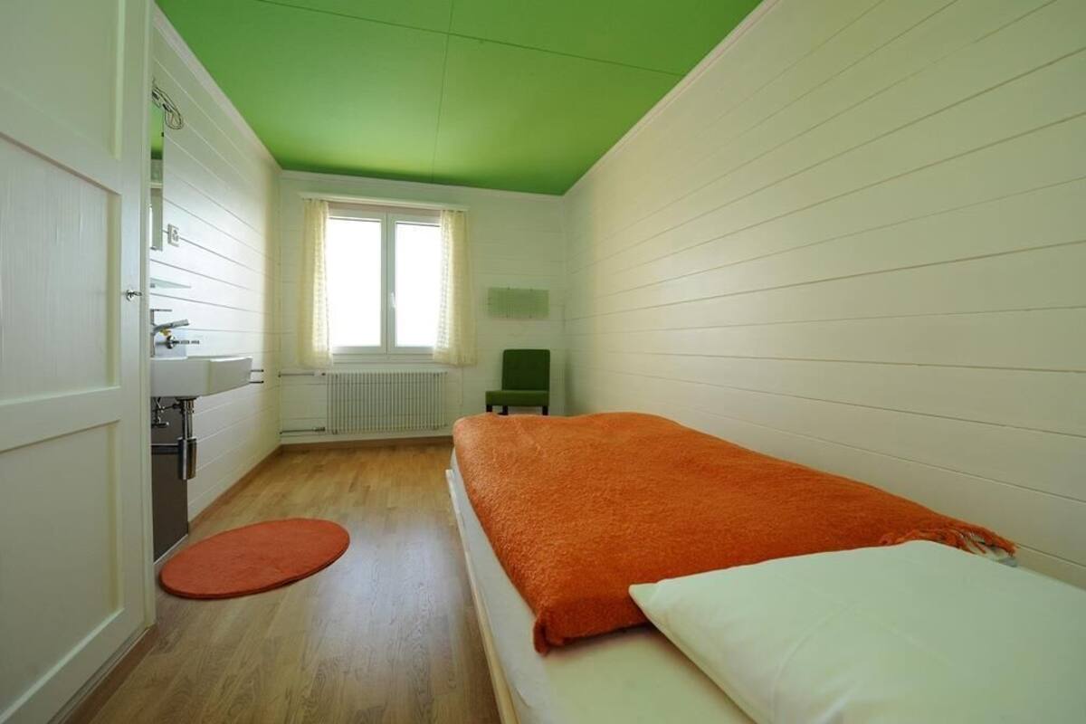 A small, single-room accommodation with a green ceiling, white walls, a wooden floor, a bed with an orange blanket, a white pillow, a small round rug, a window with curtains, a radiator, and a green chair.