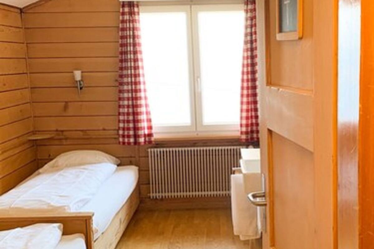 A small, cozy room with wooden walls and a sloped ceiling. There are two beds with white linens, a radiator, and a window with red and white checkered curtains.
