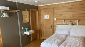 Un dormitorio con cama, mesita de noche blanca, armario de madera, espejo y un baño pequeño con lavamanos y perchero.