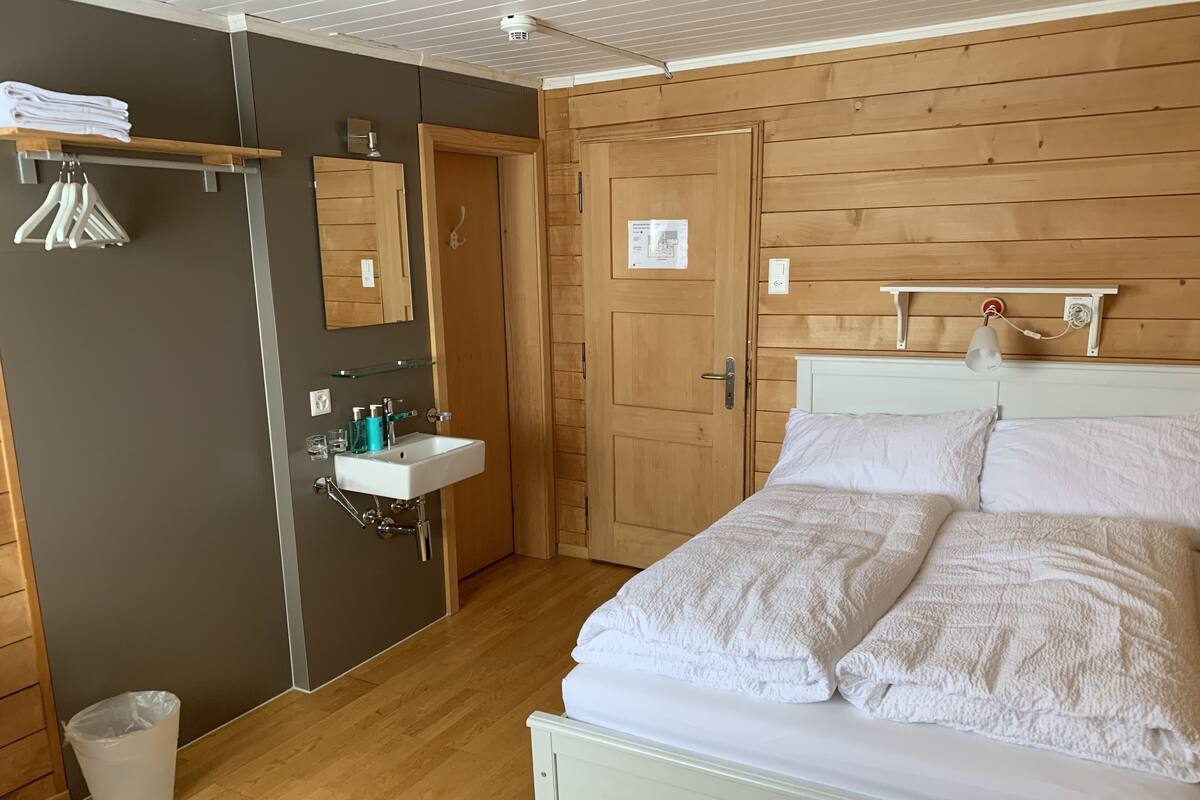 A bedroom with a bed, a white bedside table, a wooden wardrobe, a mirror, and a small bathroom with a sink and towel rack.