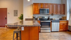 A kitchen with wooden cabinets, a granite countertop, stainless steel appliances, and a small island with stools. - Sosuite | 1BR Apt w W/D, Gym, On-Site Restaurant (Philadelphia)