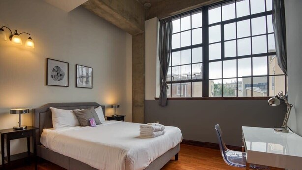 A bedroom with a bed, bedside tables, a desk, a chair, and a large window. - Sosuite | 2BR Apt w. W/D, Gym, On-Site Restaurant (Philadelphia)