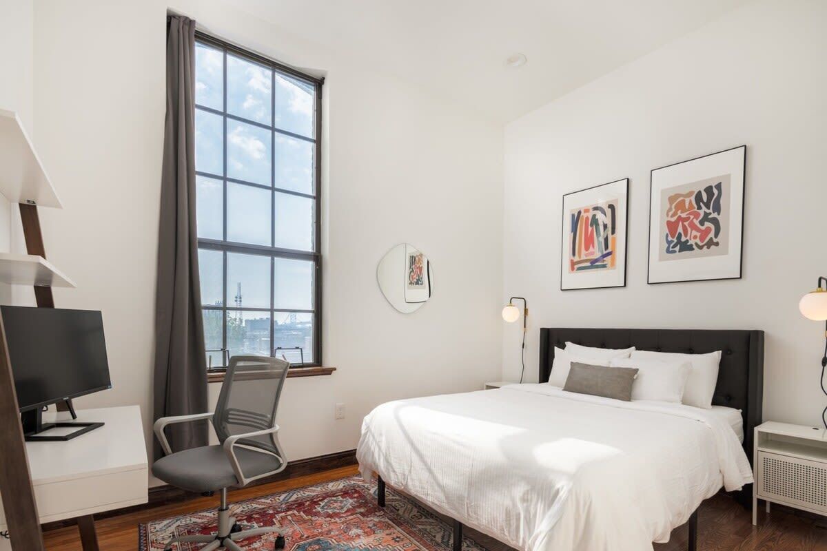 Une chambre à coucher avec un lit, un bureau avec une télévision, une chaise, une fenêtre et des œuvres d’art accrochées au mur.
