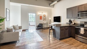 A modern kitchen with dark cabinets, a stainless steel oven, and a large island. There is a sofa and a coffee table in the living area, and a chair near the window. - Sosuite | 2BR Apt w Balcony, W/D, Gym, On-Site Bar (Philadelphia)