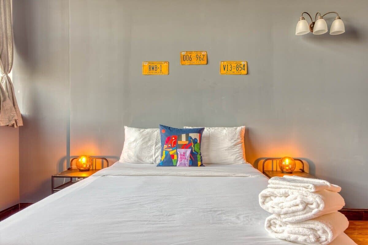 A hotel room with a bed, pillows, a colorful pillow, a stack of towels, and three wall-mounted light fixtures.