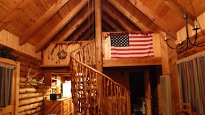 A log cabin interior with a wooden staircase, a dining area, a kitchen with a refrigerator, and a large American flag. - Circle Pine Ranch-"The Real Log Cabin Experience" (Alpine)