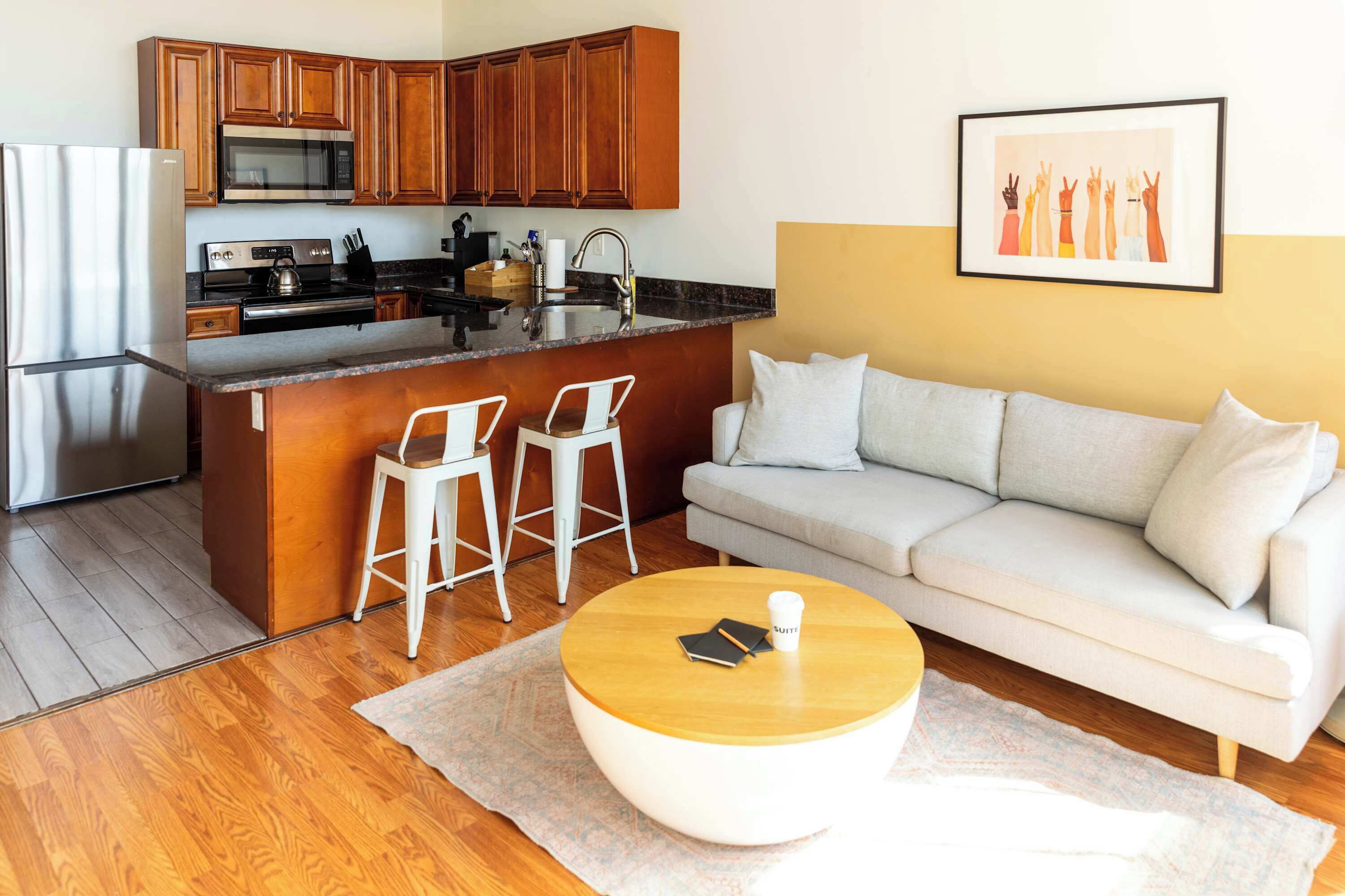 A compact kitchen with wooden cabinets, a stainless steel refrigerator, and a microwave. There is a small island with two white bar stools. A beige sofa and a round coffee table with a book and a cup are in the living area.