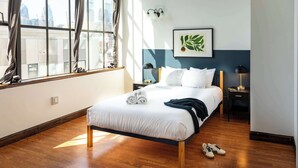 A hotel room with a large window, a bed with white bedding, a side table with a lamp, and a framed artwork on the wall. - Sosuite | 2BR Apt w W/D, Gym, On-Site Restaurant (Philadelphia)