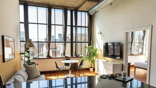 A modern living room with a large window, a flat-screen TV, a dining area, and a sofa. - Sosuite | 2BR Apt w W/D, Gym, On-Site Restaurant (Philadelphia)