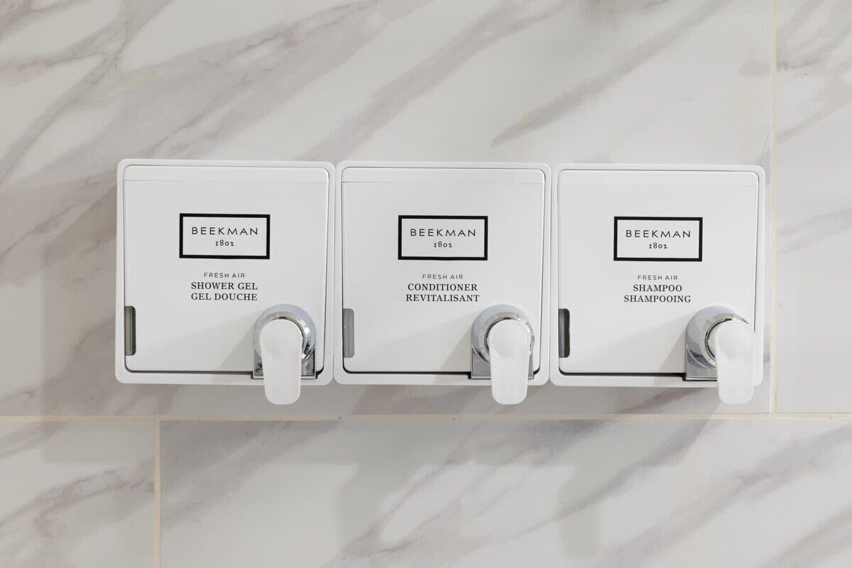 Tres dispensadores blancos con la etiqueta 'BEEKMAN', mostrando diferentes productos: 'FRESH AIR SHOWER GEL', 'FRESH AIR CONDITIONER REVITALISANT' y 'FRESH AIR SHAMPOO SHAMPOOING'.