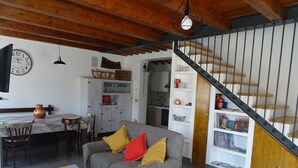 Uma sala de estar acolhedora com um sofá cinzento, tapete colorido, vigas de madeira no teto e uma escada com corrimão de madeira.