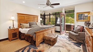 A bedroom with a large bed, wooden headboard, bedside tables, a standing fan, a window with a view of greenery, and a hot tub outside. - Enclave 158 4 Bedroom Home by RedAwning (Avon)