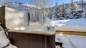 A hot tub partially covered by snow, with a clear view of a snowy landscape and trees in the background. - Enclave 158 4 Bedroom Home by RedAwning (Avon)