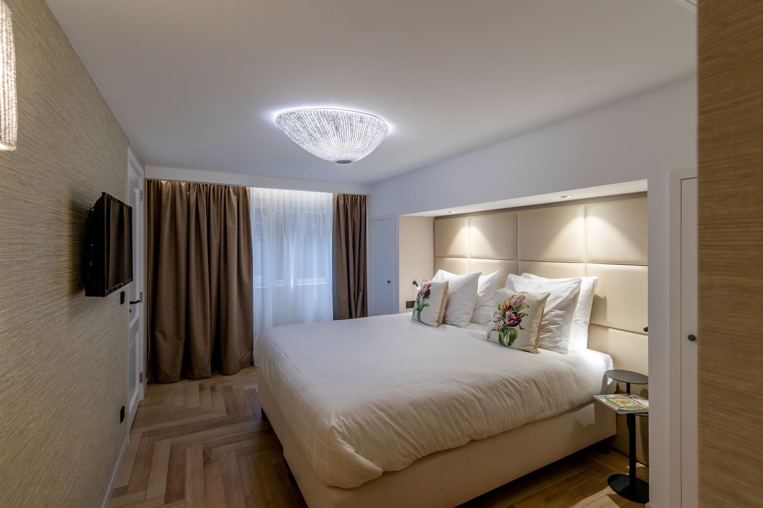 Een hotelkamer met een groot bed, een aan de muur bevestigde televisie, een kroonluchter en een tafeltje met een tijdschrift.