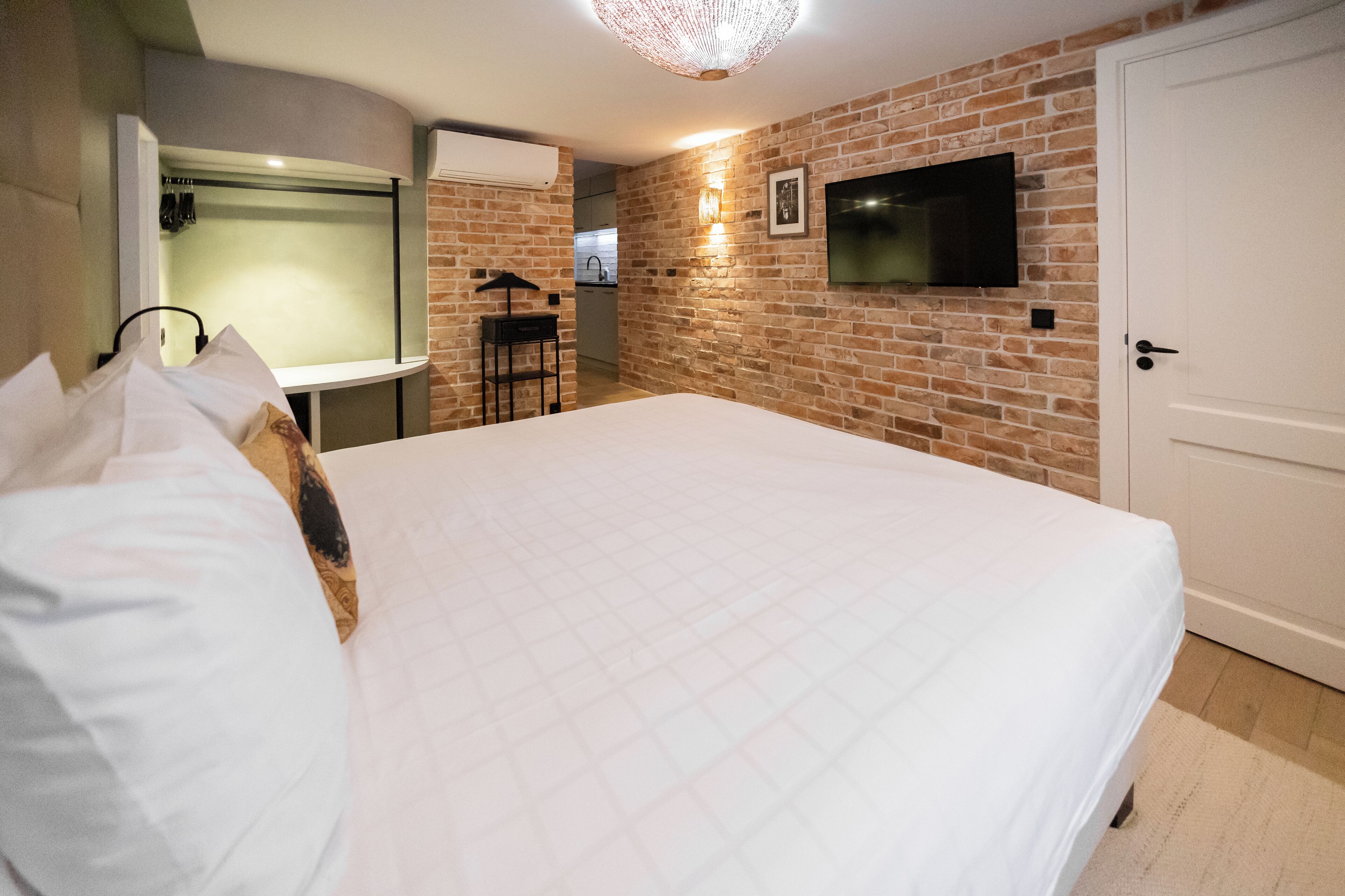 Quarto com cama grande, televisão montada na parede de tijolos e porta à direita.