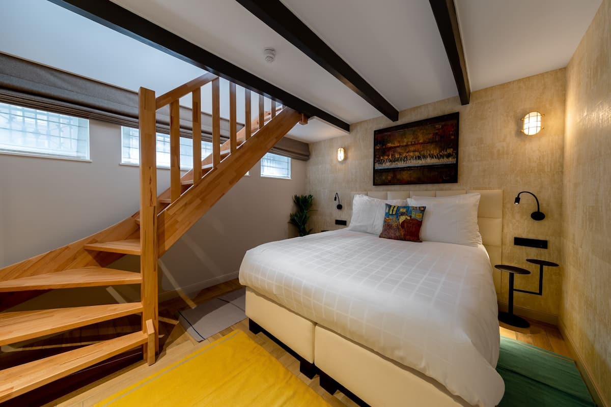 Een slaapkamer met een houten trap, een groot bed, een schilderij aan de muur en een nachtkastje met een lamp.