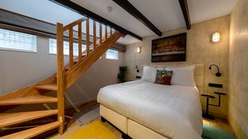A bedroom with a wooden staircase, a large bed, a painting on the wall, and a nightstand with a lamp.
