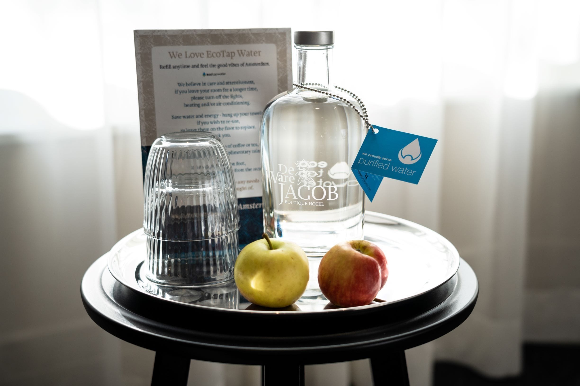 Una mesa con una botella de vidrio, dos manzanas y una jarra de vidrio. Un letrero al fondo menciona EcoTap Water.