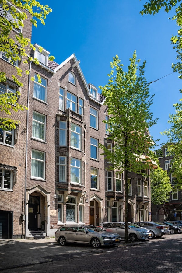 A row of brick townhouses with a tree in the foreground, parked cars along the street, and a clear blue sky. - De Ware Jacob Boutique Hotel (Amsterdam)