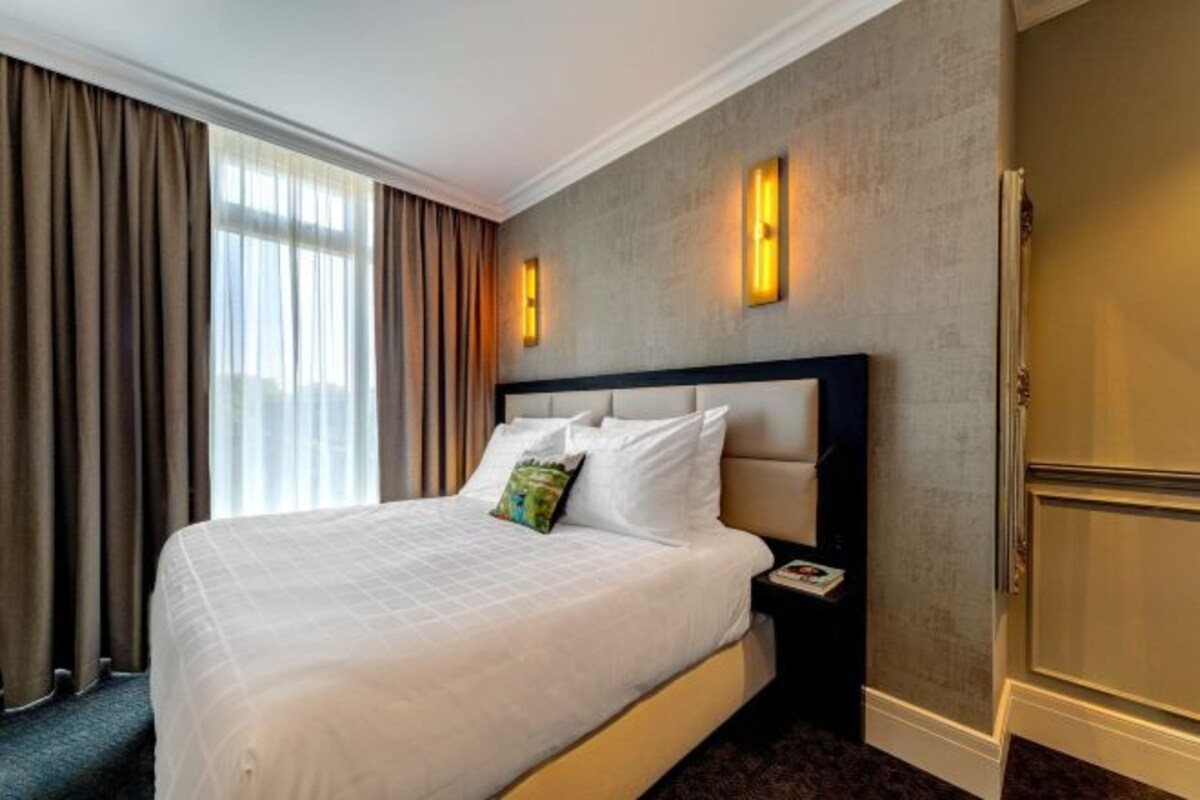 A hotel room with a large bed, a nightstand, a wall-mounted lamp, and a door.