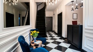 Een hotel lobby met een zwart-wit geruite vloer, een trap, een receptiebalie en blauwe stoelen met een vaas gele bloemen.