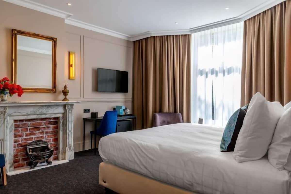Een hotelkamer met een bed, een bureau, een stoel, een open haard, een televisie en gordijnen.