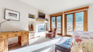 Un dormitorio con cama, escritorio, silla, chimenea y ventanas amplias con vistas a árboles.