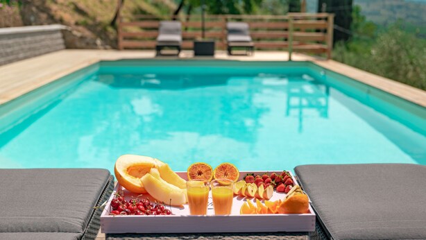 Escena junto a la piscina con una bandeja de fruta y jugo, sillas de descanso y una valla de madera al fondo.