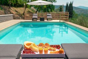 A poolside setting with a tray of fruit and juice, lounge chairs, and a wooden fence in the background.
